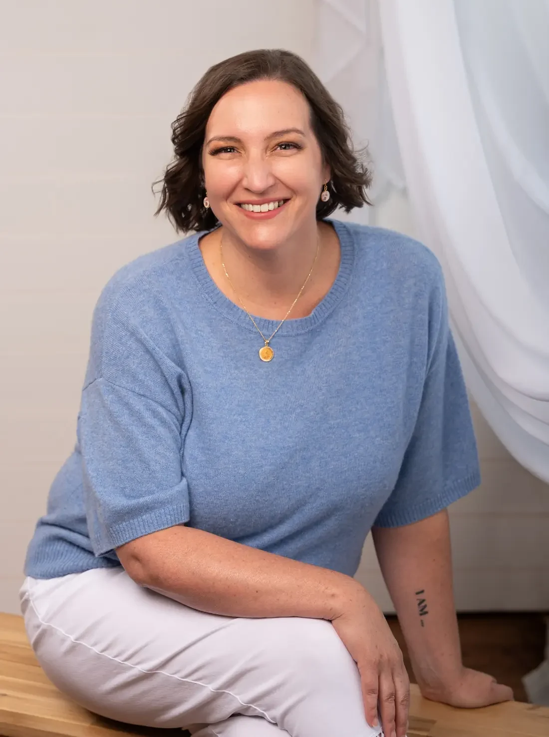Katie Quinn smiling warmly, seated on a wooden bench in a light blue sweater and white pants, with soft white drapery behind her