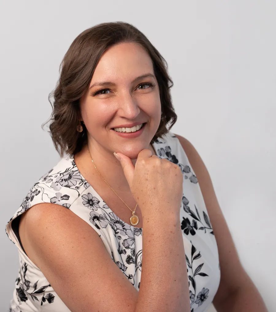 Portrait of Katie Quinn smiling warmly, chin resting on her hand, wearing a black and white floral top against a soft gray backdrop